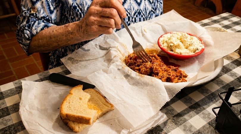 An elderly person eating pulled pork with a side of potato salad and white bread on a checkered tablecloth.