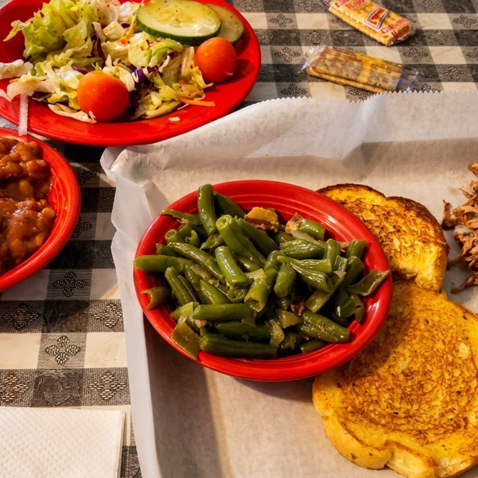 Barbecue pulled pork with toast, green beans, baked beans, and a side salad on a checkered tablecloth.