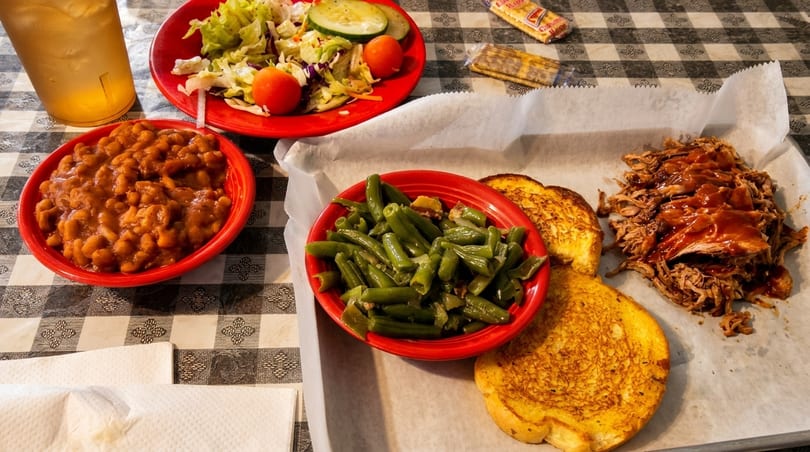 Barbecue pulled pork with toast, green beans, baked beans, and a side salad on a checkered tablecloth.
