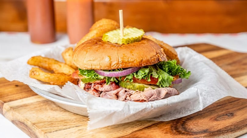 Roast beef sandwich with lettuce, tomato, onion, and pickles, served with fries on a wooden board.