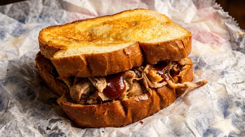 Pulled pork sandwich with barbecue sauce on toasted bread, served on crumpled white paper.