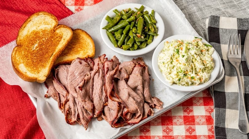 Sliced roast beef, green beans, coleslaw, and toasted bread on a tray with checkered tablecloths.